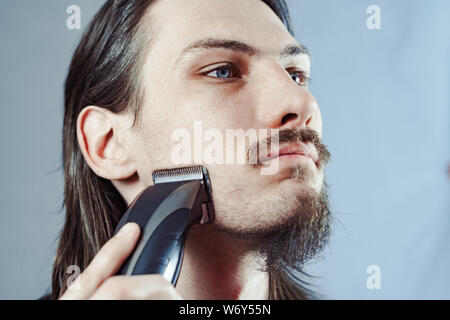 A young man shaves in the bathroom. Guy shaves his beard with a trimmer. Stock Photo