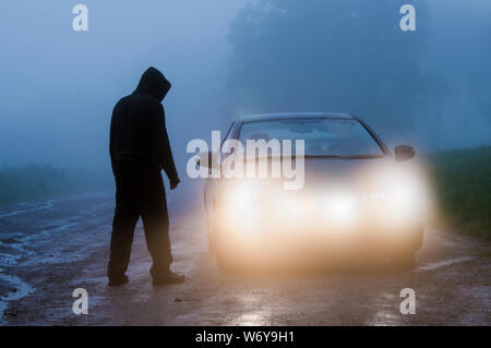 A threatening lone hooded figure standing next to a car on a misty spooky road at night. Highlighted with car headlights. Stock Photo