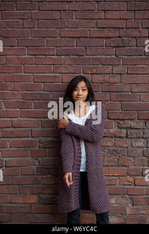 beautiful Asian young woman standing in a knitted purple warm cardigan, hugging herself over the shoulder with one hand standing against a brick wall Stock Photo