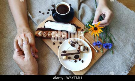 Top view of female and male hands holding each other with summer flowers cornflower and rutbeckia near a cup of black coffee and served eclairs, the c Stock Photo