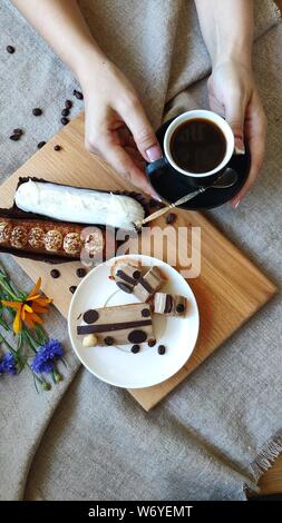 Top view on female hands with summer flowers cornflower and rutbeckia near cup of black coffee and served eclairs, the concept of a pleasant breakfast Stock Photo