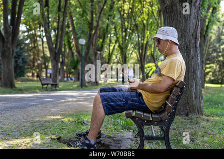 Man in hat and sunglasses listens to music sitting down on bench in park. Man listens to music with phone while relaxing in the park under trees Stock Photo