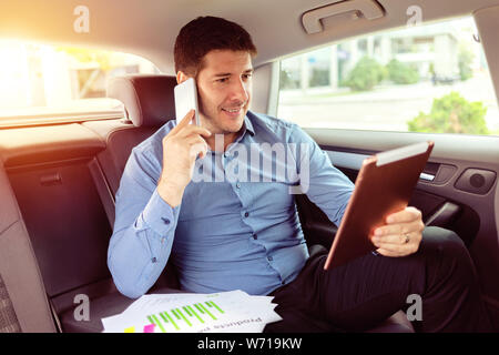 Successful smiling businessman talking on phone while sitting in back seat of car using digital tablet while commuting Stock Photo