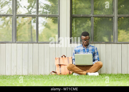 Full length portrait of smart African student using laptop outdoors while sitting on green grass in college campus, copy space Stock Photo