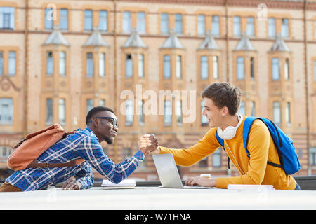 Side view portrait of two contemporary college students, one of them African, shaking hands while standing on opposite sides of table outdoors Stock Photo