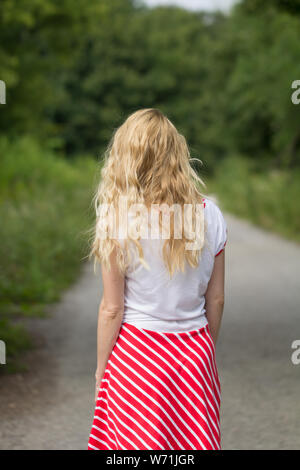 rear view of blonde woman wearing red skirt walking on forest path Stock Photo