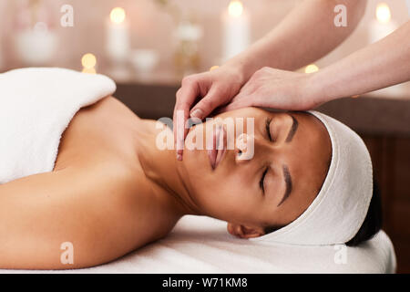 Portrait of young mixed-race woman enjoying face massage in luxury spa, copy space Stock Photo