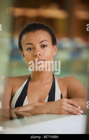Portrait of gorgeous mixed race woman relaxing in swimming pool and looking at camera Stock Photo