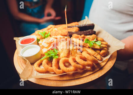 Assorted beer snacks: onion rings, cheese balls, chips etc on a iron tray on a dark wooden background among vegetables. Close-up. Space. Stock Photo
