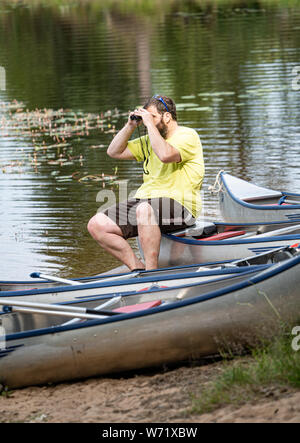 Man looking through binoculars, sitting on a canoe. Black River (Svartälven) in the wilderness, Sweden Stock Photo