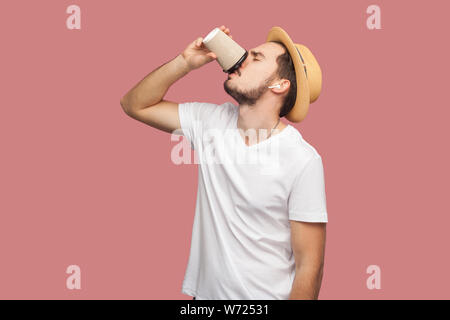 Portrait of handsome bearded young hipster man in white shirt and casual hat standing and drinking coffee in paper cup, freshness lifestyle. Indoor, i Stock Photo