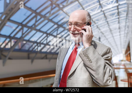 Senior business man dressed in suit and eyeglasses is talking on the mobile phone and smiling while sitting in modern building Stock Photo