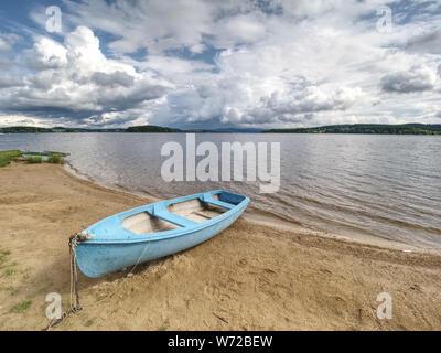 Blue fishing boat anchored on beach sand of lake. Smooth evening water level and calm. Large vallely of Lipno lake. Stock Photo