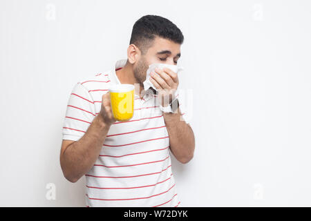 Cold and flu. Portrait of sick bearded young man in striped t-shirt standing, holding tissue, cleaning his nose, holding hot drink, try to be treated. Stock Photo
