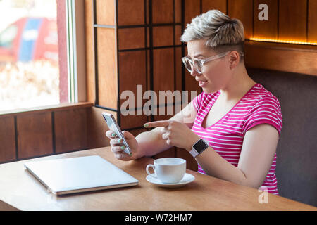 Side view portrait of excited young blogger with short hair in pink t-shirt is sitting in cafe, holding phone and making video call with shocked face, Stock Photo