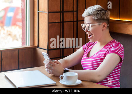 Side view portrait of shocked screaming young blogger with blonde short hair in pink t-shirt is sitting in cafe, holding phone with opened mouth, read Stock Photo