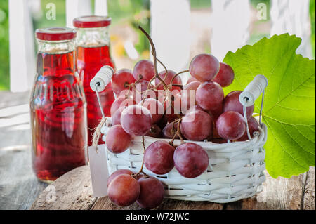 A bunch of pink grapes, prepared to extract the juice, is in a white basket . Two bottles of grape juice are on the table next to a bunch of grapes Stock Photo