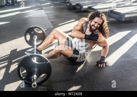 Portrait of grimace young adult man athlete with long curly hair working out in gym, sitting on floor and have strong hurt problem with shelder, spasm Stock Photo