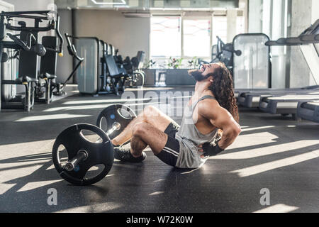 Portrait of injury young adult man athlete with long curly hair working out in gym, sitting on floor and have strong hurt problem with back, spasm pai Stock Photo