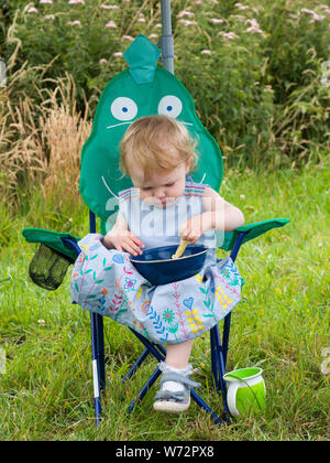 Toddler sat on a child size garden chair having a picnic lunch, UK