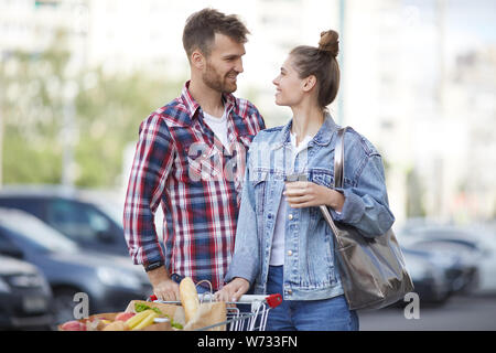 Waist up portrait of contemporary young couple pushing shopping cart with groceries in parking lot, copy space Stock Photo