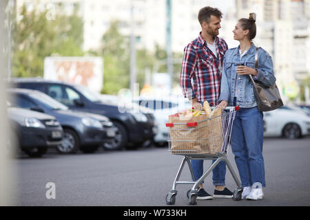 Full length portrait of contemporary young couple pushing shopping cart with groceries in parking lot, copy space Stock Photo