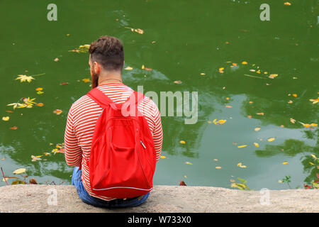 Young man tourist with a red backpack sits on a lake on a warm autumn day. Yellow fallen leaves float on the water in leaf fall. Stock Photo