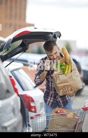 Portrait of handsome young man speaking by phone while packing groceries into car trunk Stock Photo