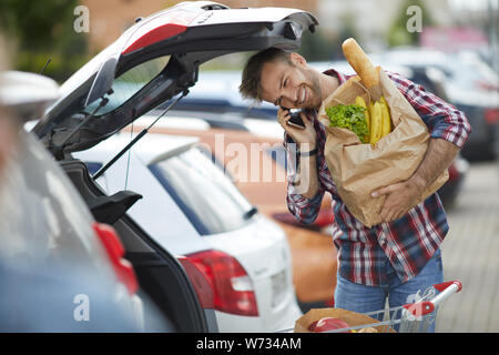 Portrait of smiling young man speaking by phone while packing groceries into car trunk , copy space Stock Photo