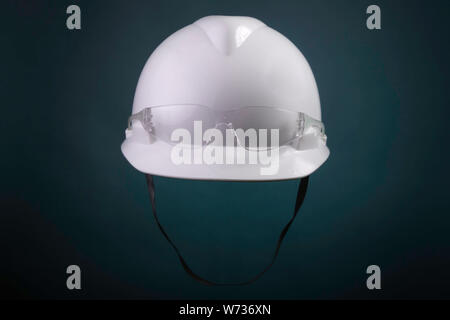 White generic protective construction helmet with strap and plastic transparent safety glasses on the cap against a green studio background Stock Photo