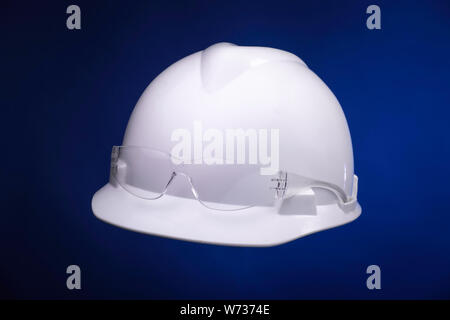 White generic protective construction helmet with a plastic transparent safety glasses on the cap against a blue studio background Stock Photo