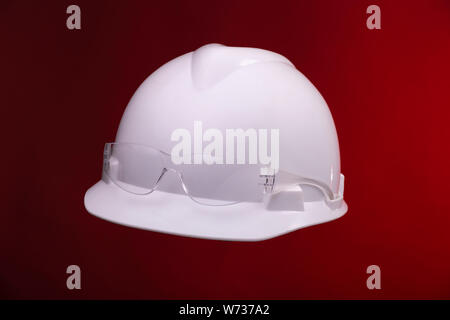 White generic protective construction helmet with a plastic transparent safety glasses on the cap against a red studio background Stock Photo