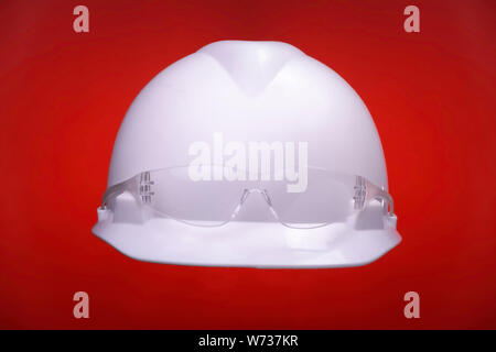 White generic protective construction helmet with a plastic transparent safety glasses on the cap against a light red studio background. Stock Photo