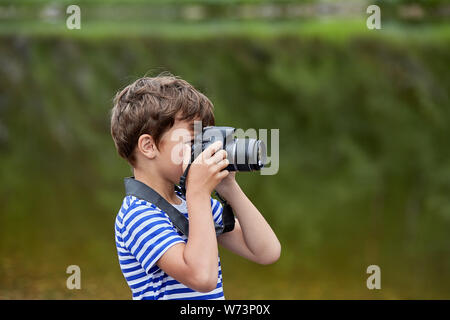 White boy about 8 years old is standing near river and taking pictures by digital camera. Stock Photo