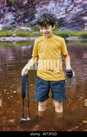 White pretty smiling boy about 8 years old is standing in river with digital camera partly in water, he is cheerful. Stock Photo