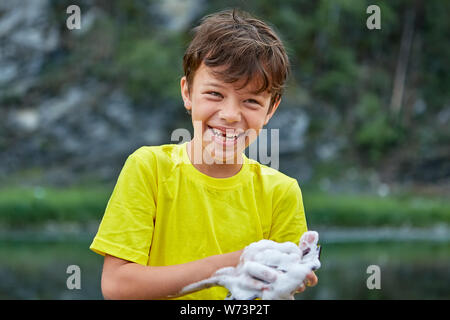 White cheerful boy about 8 years old is standing in river's water and washing digital camera using foam of soap, he is smiling. Stock Photo