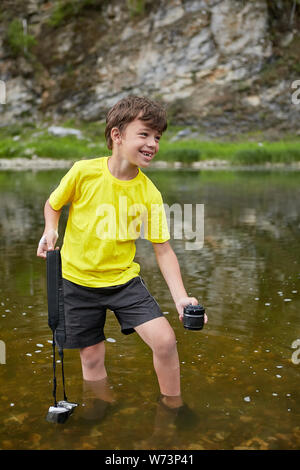 White cheerful boy about 8 years old is watering digital camera in river, his legs and camera are in water. Stock Photo
