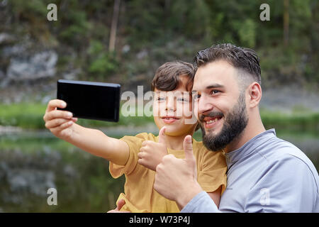Father and son are using smartphone for taking pictures of themselves in forest near river, eco-tourism. Stock Photo