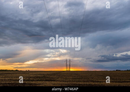 A row of high voltage electrical power lines cling to the horizon in an otherwise vast, wide open rural landscape on sunset with clouds. Stock Photo