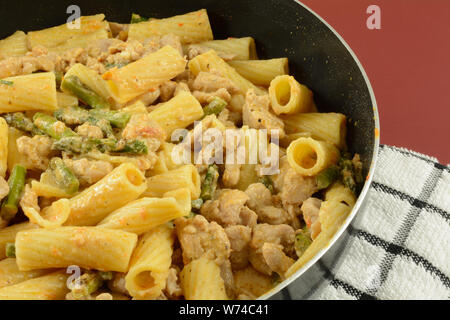 Hot rigatoni pasta with chicken and asparagus cream sauce in non stick skillet on towel on red table Stock Photo