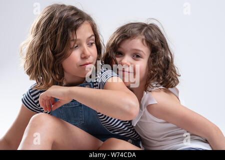 Two attractive young sisters cuddling together with the younger little girl giving the camera a happy friendly smile in a close up portrait over a whi Stock Photo