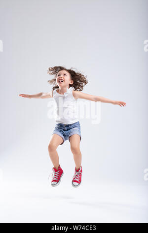 Joyful energetic active little girl in denim shorts and red sneakers jumping high into the air with outstretched arms over a white background with cop Stock Photo