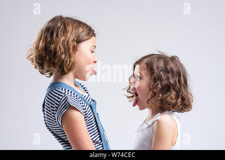 Mischievous naughty little sisters sticking out their tongues at each other as they face off in a side view portrait isolated on white Stock Photo