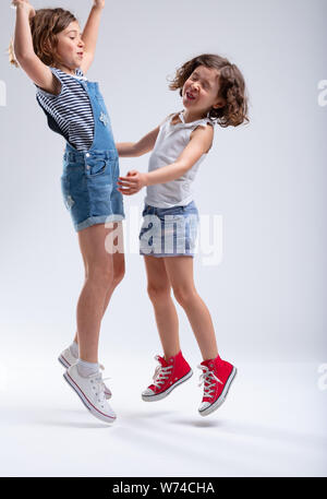 Two young sisters jumping and laughing together in blue denim shorts as they enjoy their summer vacation over a white background Stock Photo