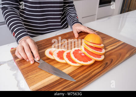 Female hands placed on wooden chopping board with sliced red grapefruit and knife Stock Photo