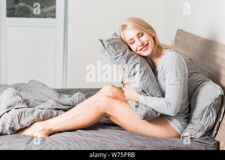 Portrait of happy female awakes in good mood after healthy dream, sitting on comfortable bed, hugging gray soft pillow in her bedroom. Relaxed beautif Stock Photo