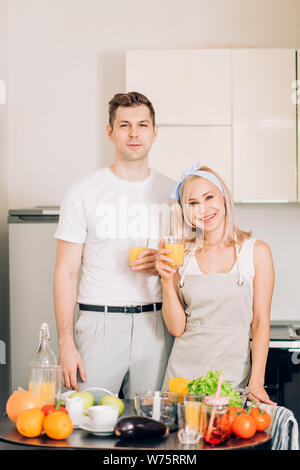 Happy young caucasian couple making organic drink together. Shot of relaxed joyful young people holding glasses with orange juice, smiling looking at Stock Photo