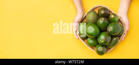 Hand holding organic avocados whole fruit in basket on yellow table background.Healthy super foods for diet.Fresh vegetable from farm.keto food ingred Stock Photo