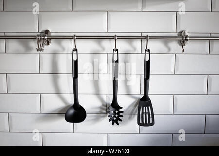 Black plastic kitchen utensil hang on white stone wall. Photo of cookware. Kitchen utensils hanging white brick background. Stock Photo