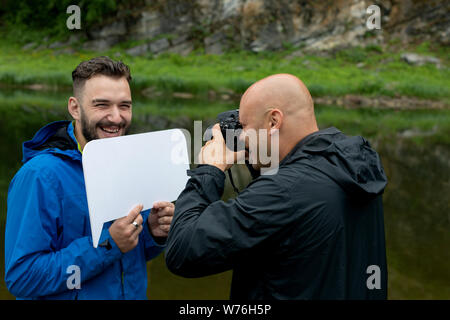 Taking a picture or a photo shoot in the open air. A bald male photographer is photographing a model of a bearded guy holding a mocap sign for text. Stock Photo